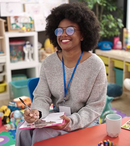 A smiling Preschool Teacher Aide with curly hair and glasses sits in a colorful classroom, holding a clipboard and pen. She wears a gray sweater and blue lanyard, with toys and drawings visible in the background.