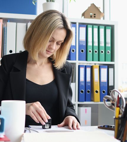 A Procurement Clerk with blonde hair wearing a black blazer is sitting at a desk, pressing a stamp onto a document. Behind her are shelves filled with colorful ring binders and files.