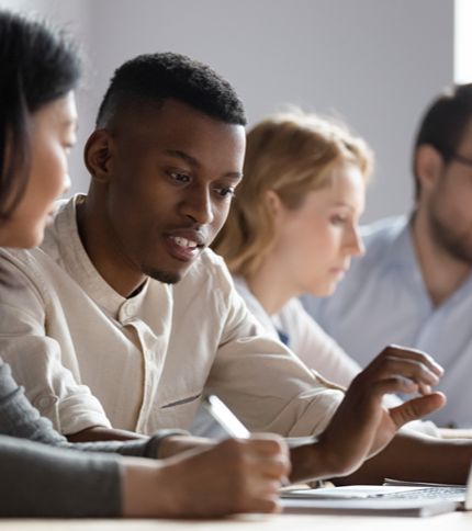 A group of four people sit side by side at a table, focused on discussion and writing. The Program Coordinator in the foreground engages in conversation, while two others appear to be working in the background.
