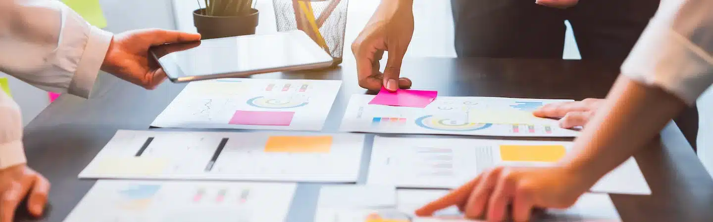 Three people collaborate on a project with charts, graphs, and notes spread out on a table and a tablet in hand.