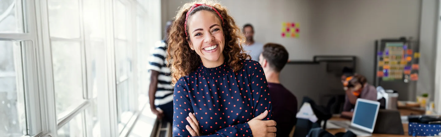Smiling woman in a polka dot shirt stands with arms crossed in a bright office with people working in the background.