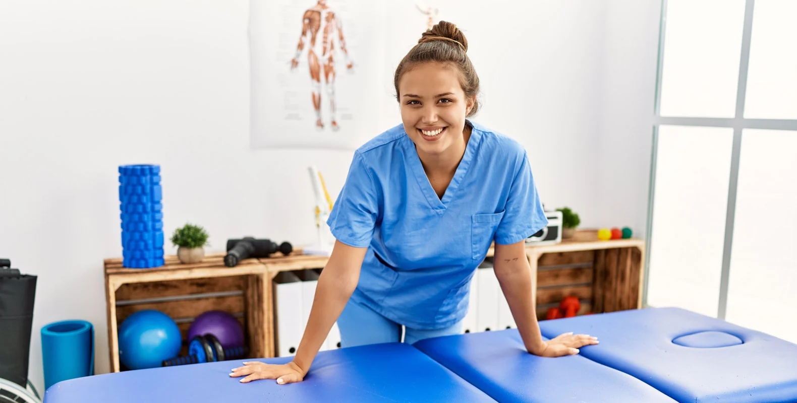 A smiling person in blue scrubs stands beside a massage table in a therapy room filled with exercise equipment.
