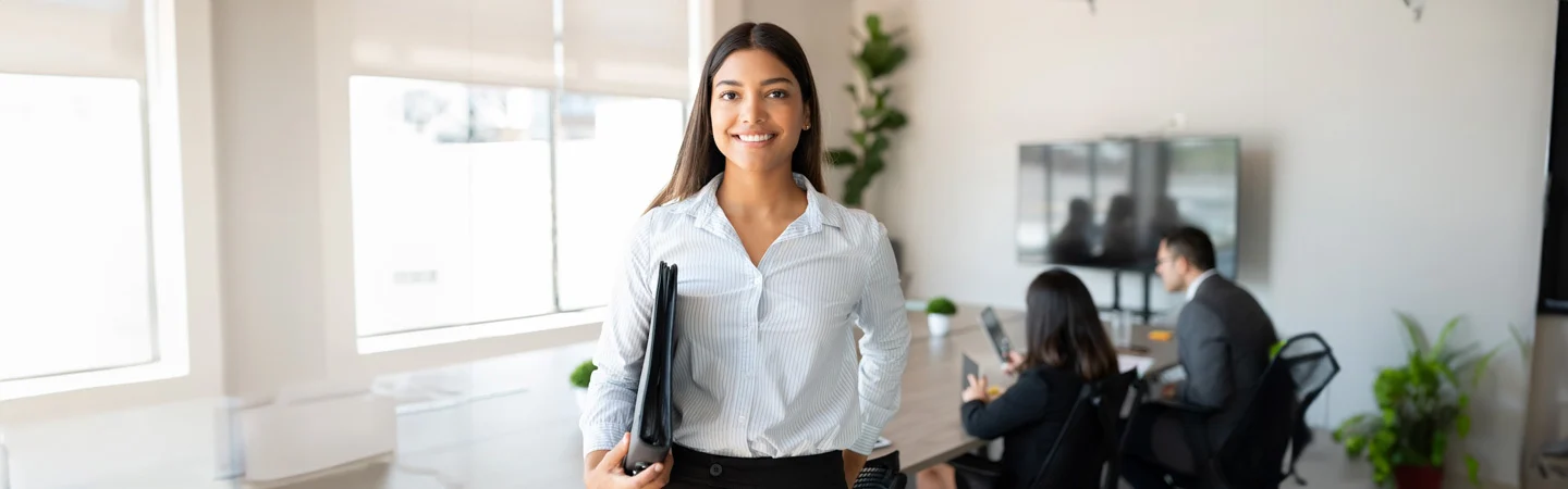 Smiling woman in business attire holding a folder, standing in a modern office with colleagues working in the background.