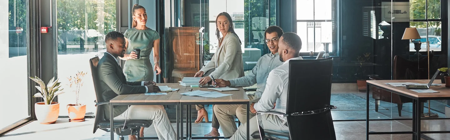 Five people having a meeting around a table in a modern office with large windows and natural light.