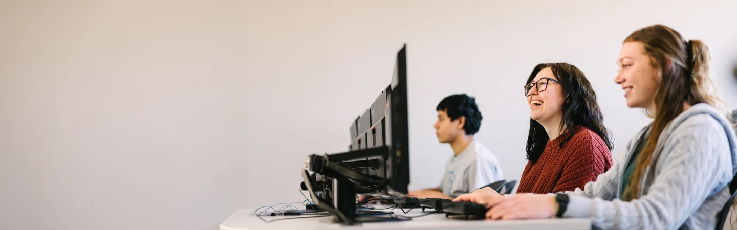 Three people sitting at desks, working on desktop computers, with a plain wall in the background.