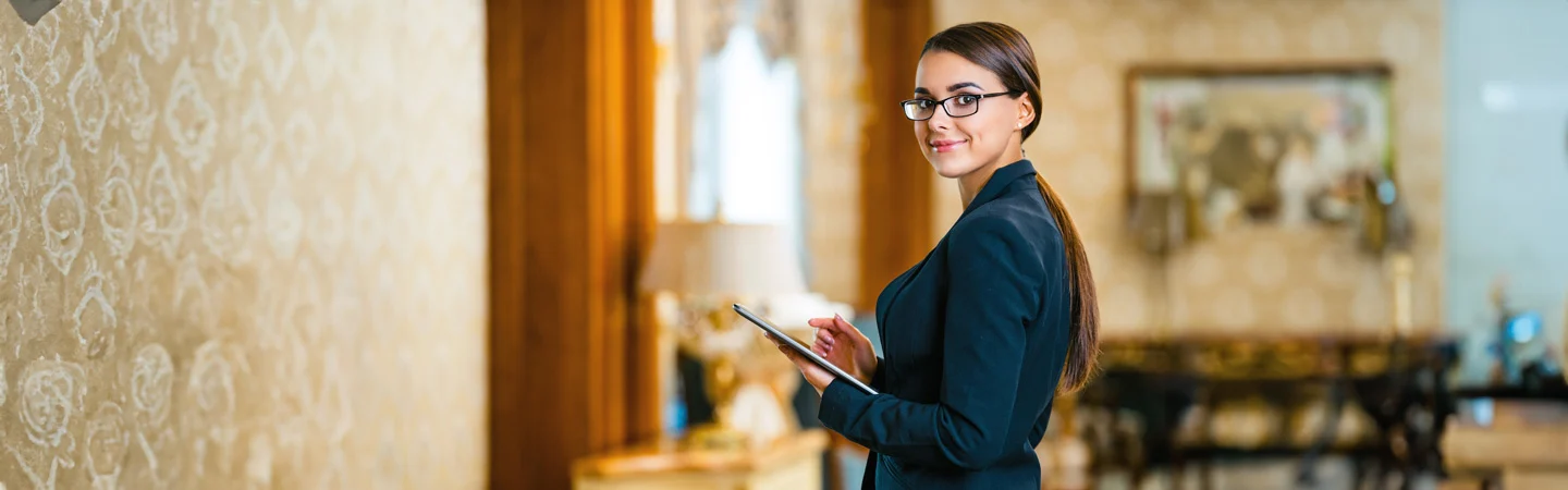 A woman in a suit and glasses holds a tablet, standing in an elegantly decorated room with ornate wallpaper.