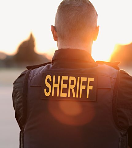 A Deputy Sheriff wearing a vest labeled “SHERIFF” stands outdoors at sunset, facing away from the camera.