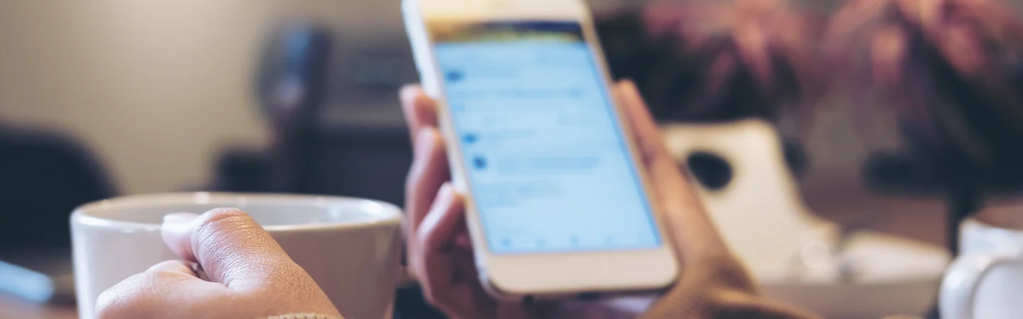 Person holding a white ceramic cup in one hand and a smartphone in the other, with a blurred background.