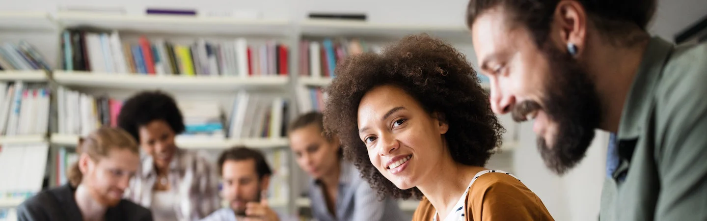 A diverse group of people working together, smiling and collaborating in a bright room with bookshelves in the background.