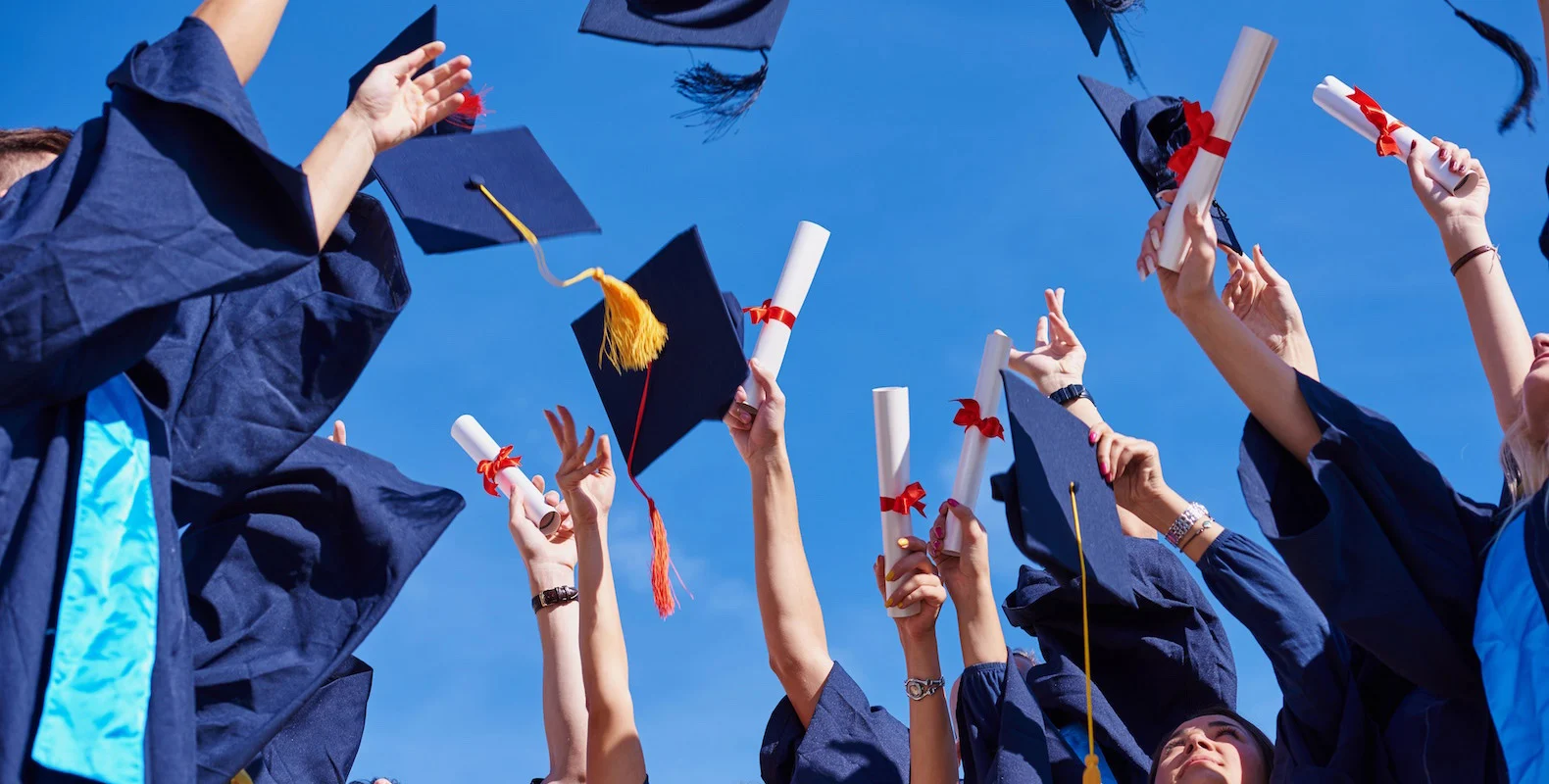 Graduates in caps and gowns joyfully toss hats and hold diplomas against a clear blue sky.