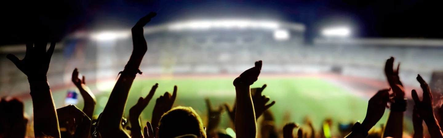 Silhouettes of cheering fans with raised hands in a stadium at night.