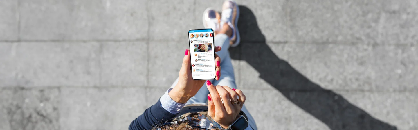 Person with red nails holds phone displaying social media feed while standing on grey pavement.