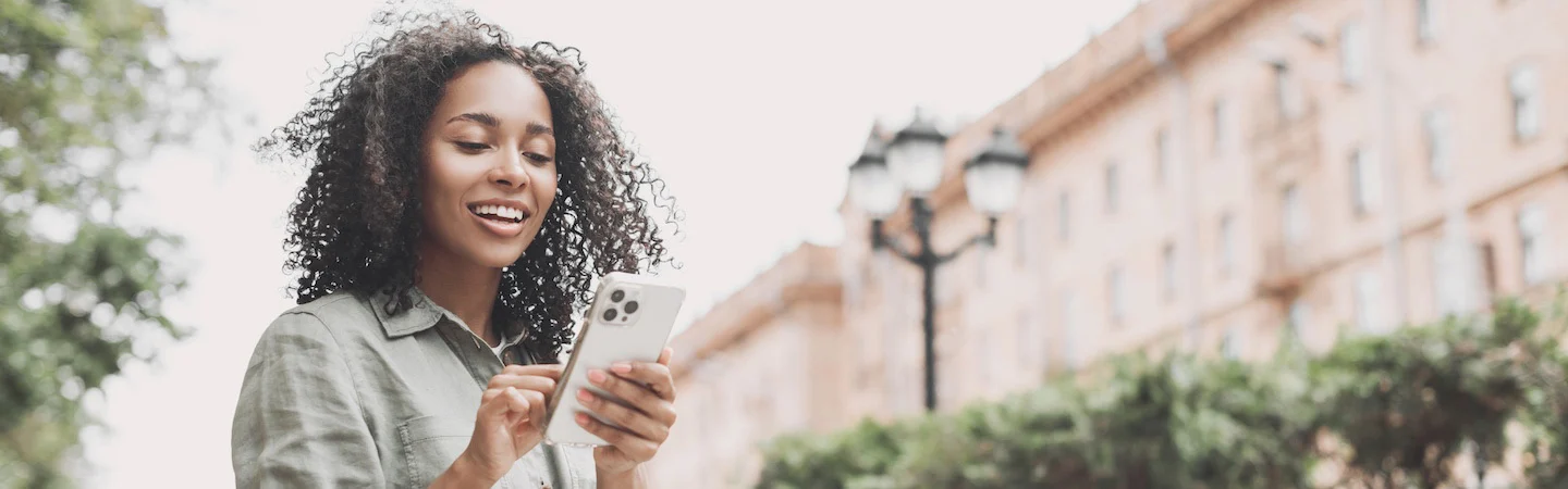 A woman stands outdoors, smiling and using her smartphone. Trees and a building with street lamps are in the background.