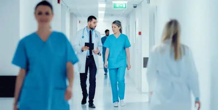Doctors and nurses walk through a bright hospital corridor, with some engaged in conversation.