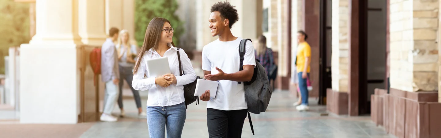 Two students walking and talking on a college campus, both holding notebooks and wearing backpacks.