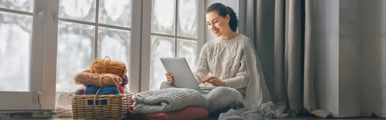 Woman in a cozy sweater sits by a window with yarn and a laptop, smiling and enjoying her time indoors.