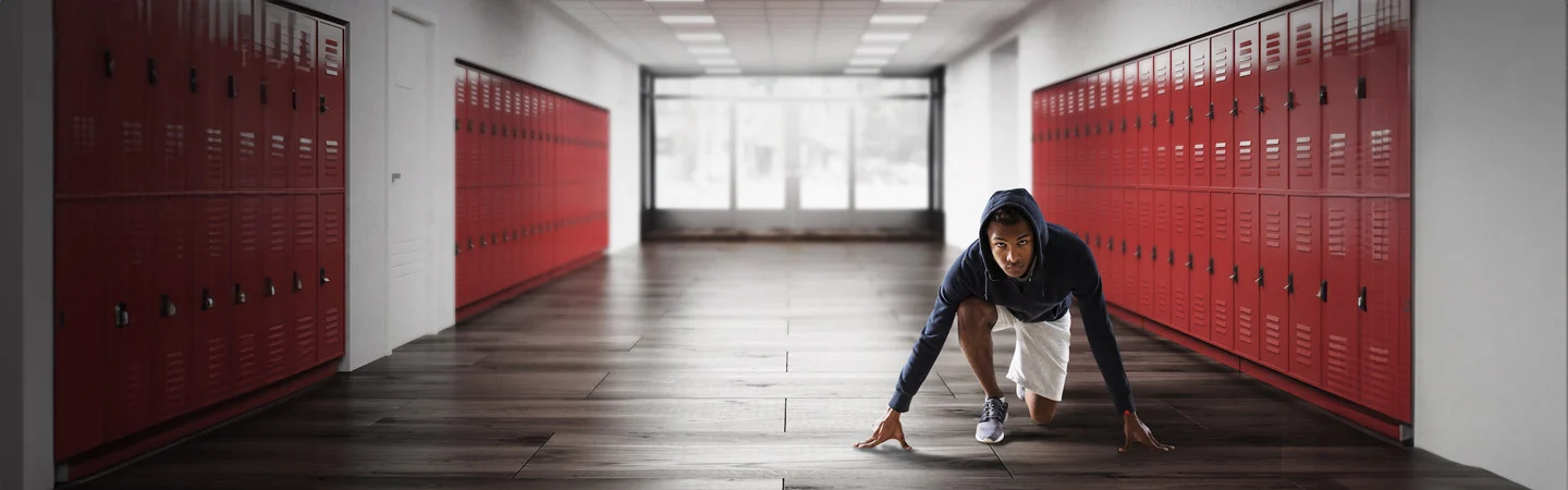 Person in a hoodie crouched in a sprinter's starting position in a school corridor with red lockers.