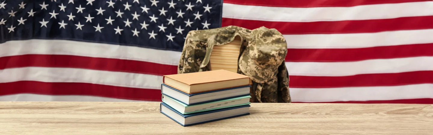 Books stacked on a table with a military uniform jacket draped over a chair and an American flag in the background.