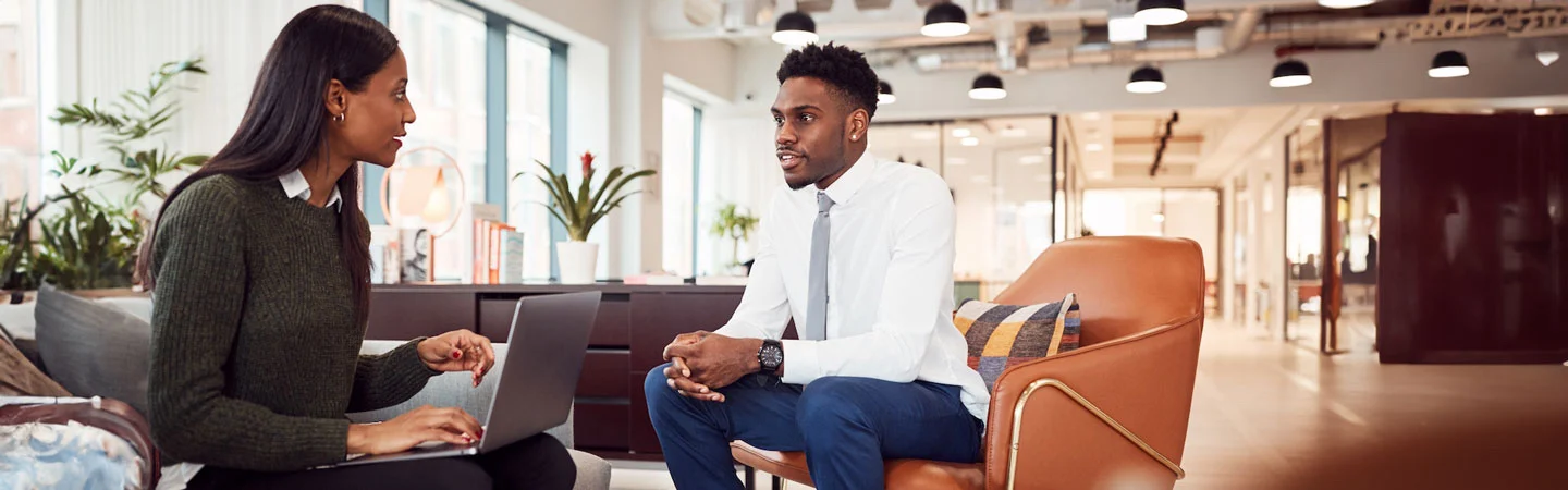 Two colleagues discuss work while seated in a modern office; one uses a laptop, the other listens attentively.