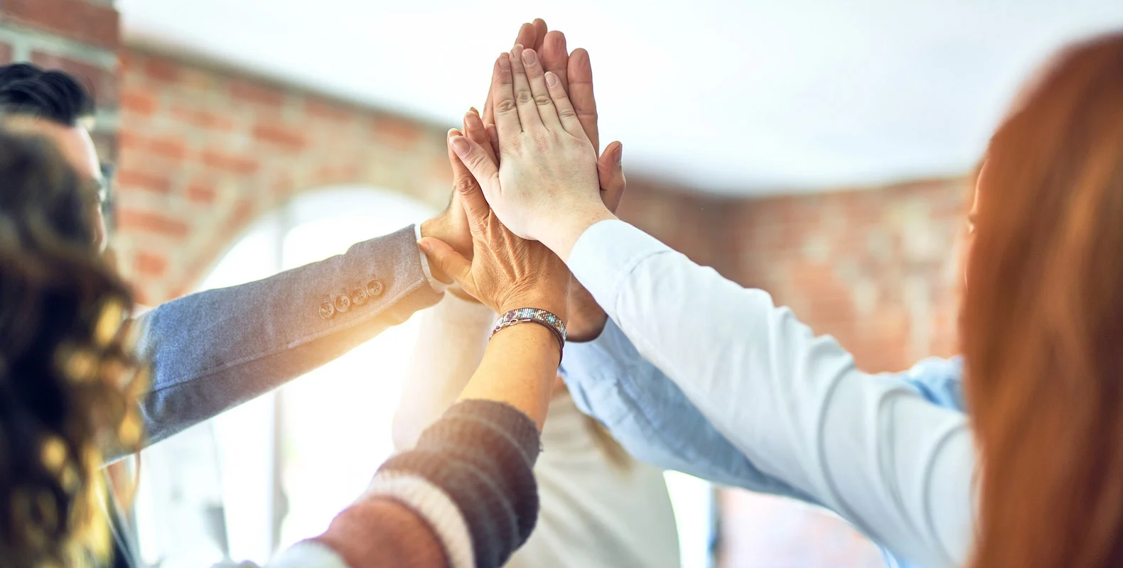A group of people join hands in a high-five, symbolizing teamwork and unity in a well-lit room with brick walls.
