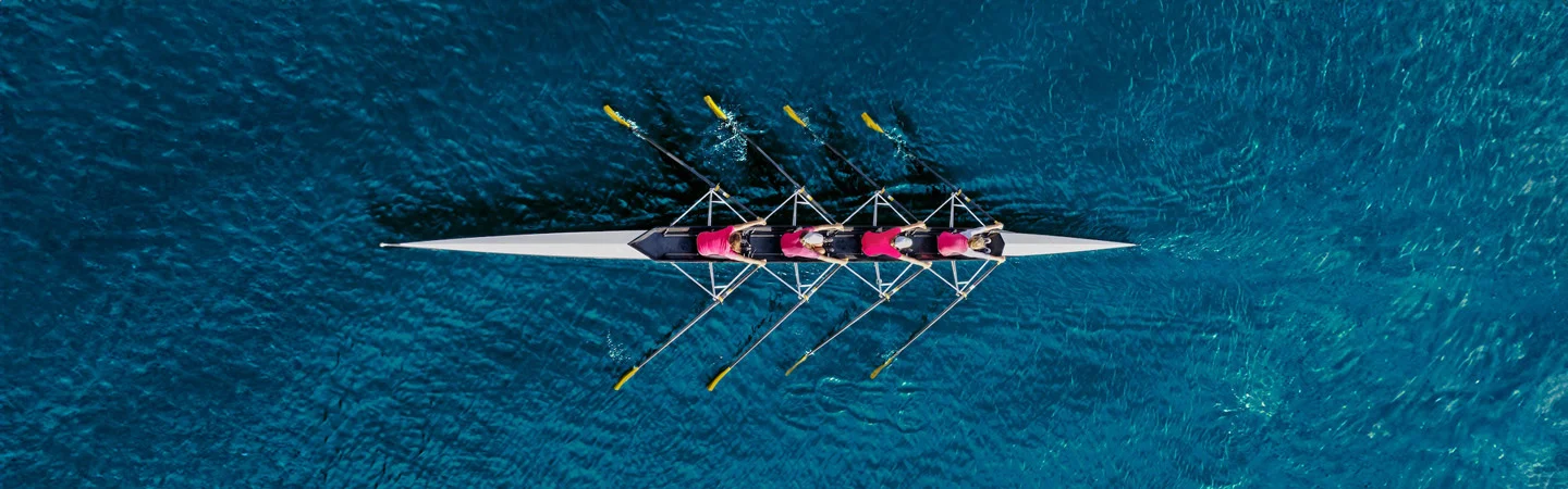 Aerial view of four rowers in a boat, rowing in sync on a deep blue body of water.