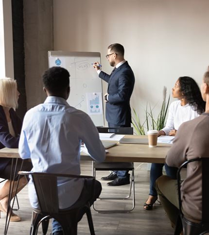 A man in a suit, likely the training coordinator, stands by a flip chart giving a development-focused presentation to four colleagues seated around a table in a modern office. Graphs and charts are visible on the flip chart.