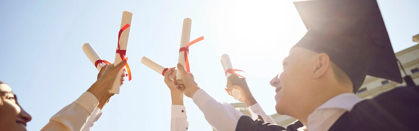 Graduates in caps and gowns hold up diplomas with red ribbons, celebrating under a sunny sky.