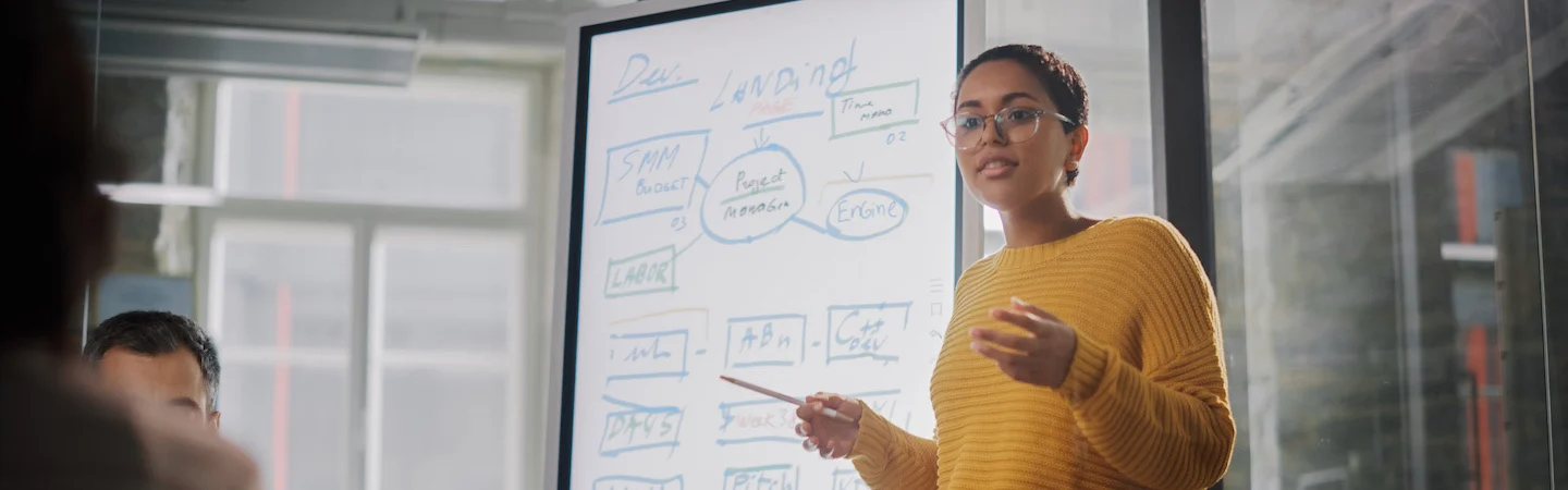 Person in a yellow sweater presenting with a pointer at a whiteboard covered in various notes and diagrams.