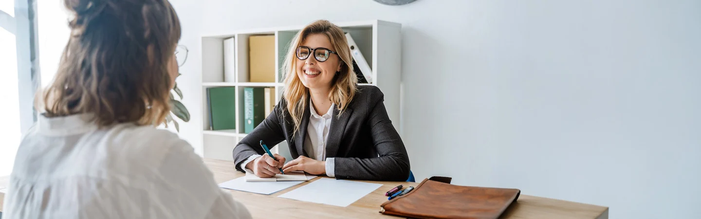 A smiling woman in glasses and a blazer sits at a desk with papers, interviewing another woman with curly hair.