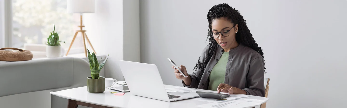 Woman working at a desk with a laptop, calculator, and papers, holding a smartphone, plants and decor around her.