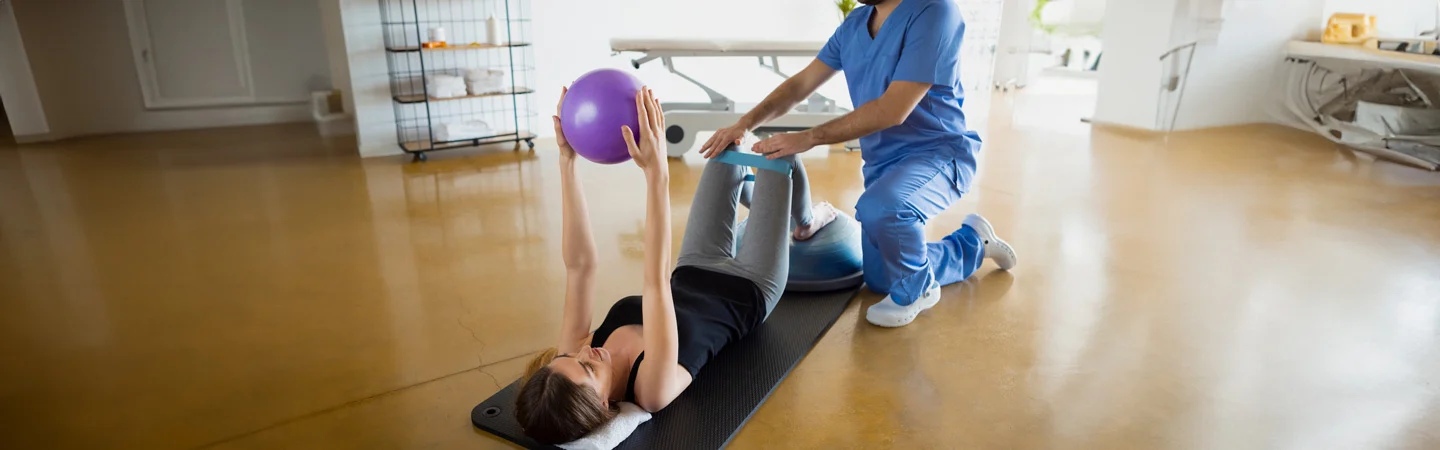 A physical therapist in blue scrubs helps a patient lying on a mat with an exercise ball in a physiotherapy session.