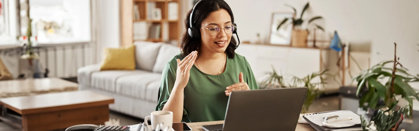 A woman wearing headphones sits at a desk in a bright room, engaged in a video call on her laptop.