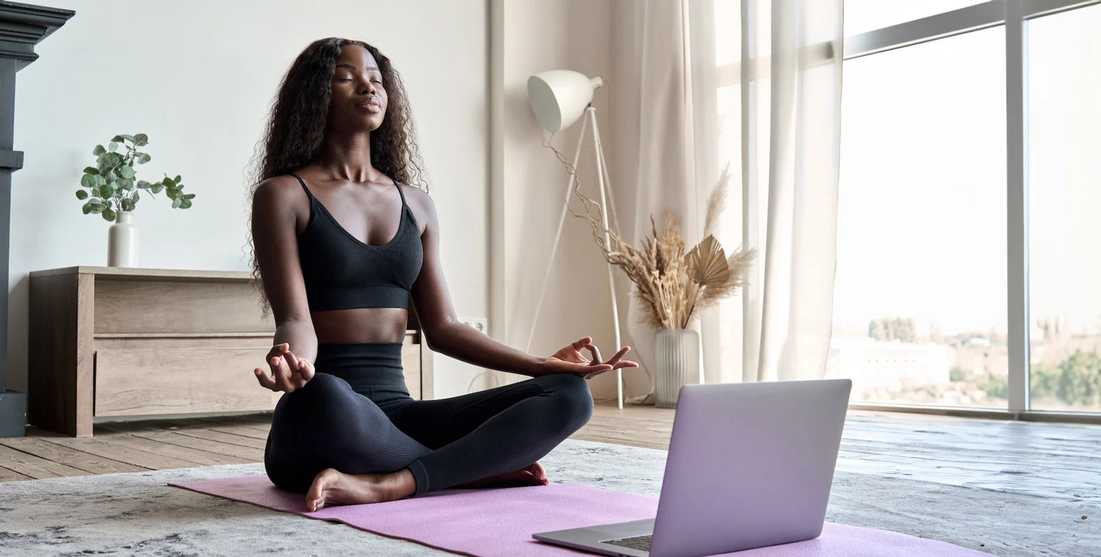 A woman in black activewear sits cross-legged on a yoga mat, meditating in front of a laptop at home.