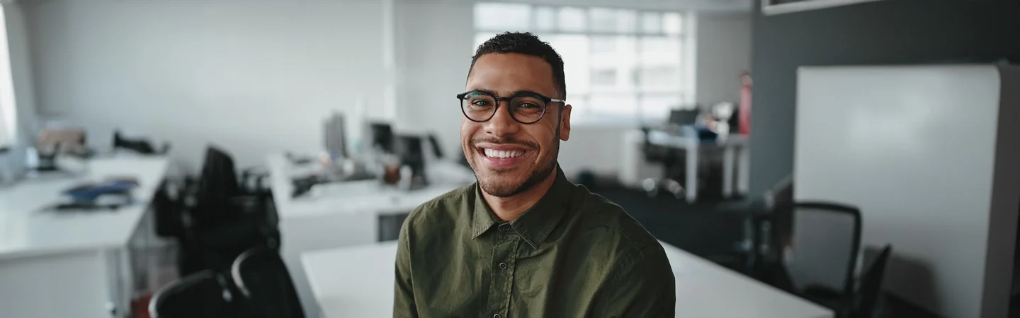 Smiling man wearing glasses and a green shirt in a modern office setting with desks and computers in the background.