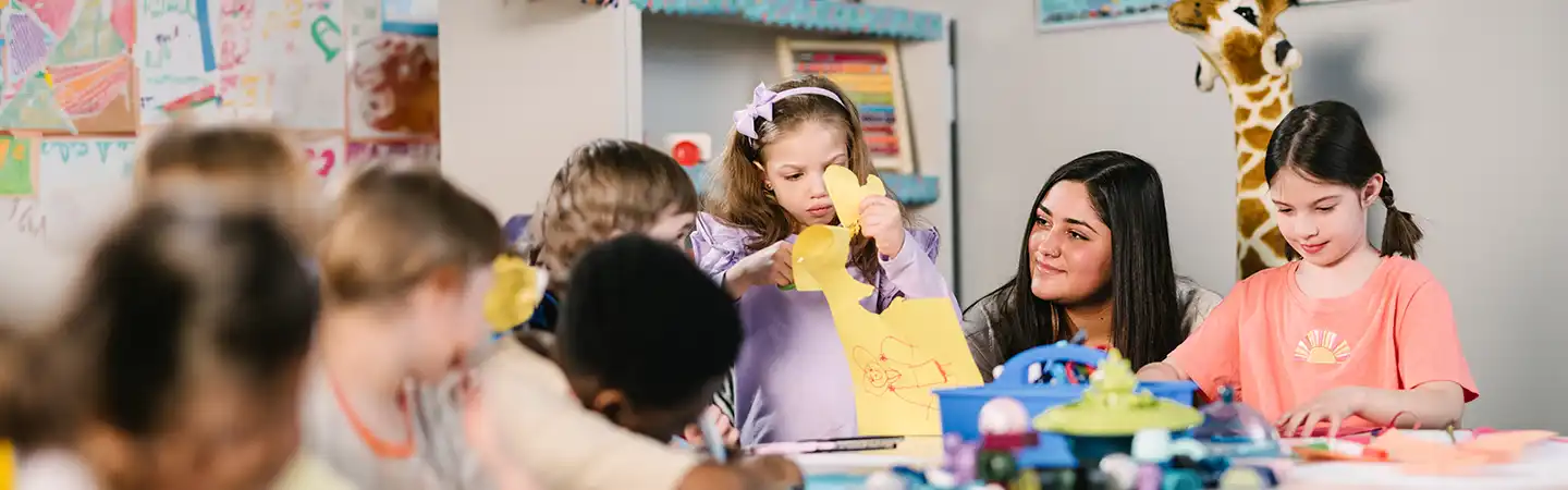 Children doing arts and crafts in a classroom, with a teacher assisting them. A giraffe toy is visible in the background.