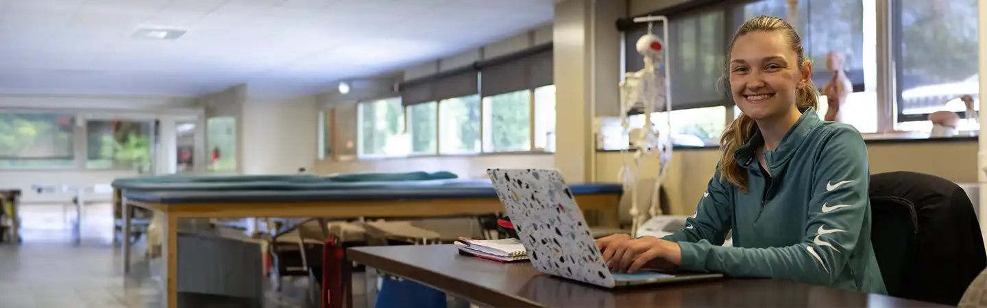 A person sitting at a desk with a laptop, smiling in a spacious, bright room with multiple windows and medical equipment.