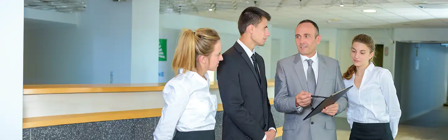 Four professionals having a discussion in an office hallway, three wear business attire, one holds a clipboard.