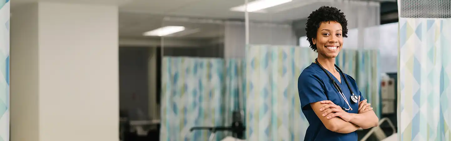 Smiling healthcare worker in blue scrubs with arms crossed, standing in a medical facility with patterned curtains in the background.