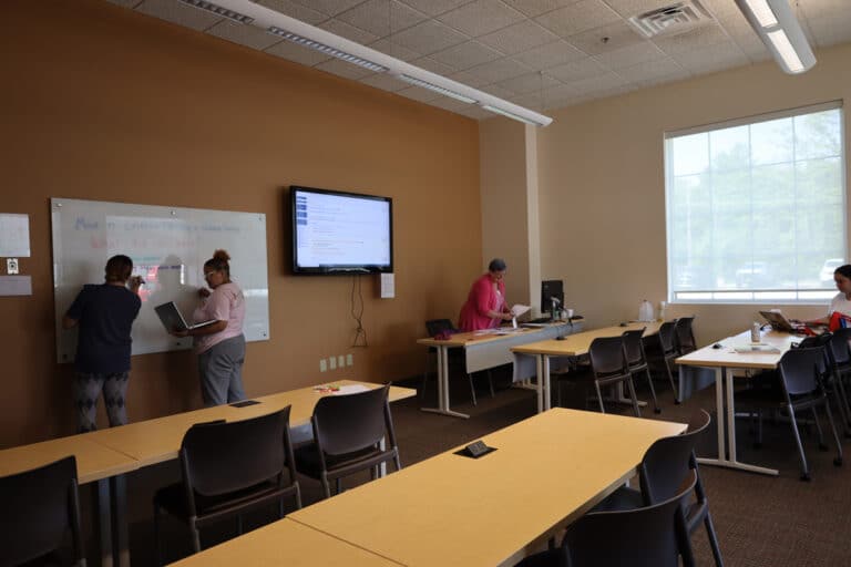 Three people working in a classroom, two writing on a whiteboard, and one using a computer.
