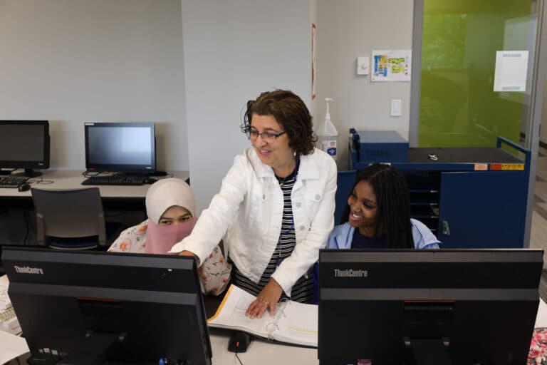 A teacher assists two students working on desktop computers in a classroom.