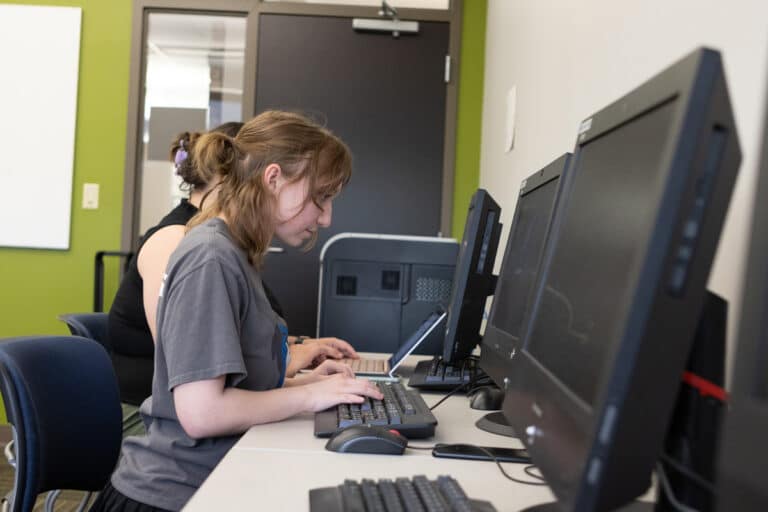 Person typing on a keyboard at a computer desk in a classroom, with another person in the background using a laptop.