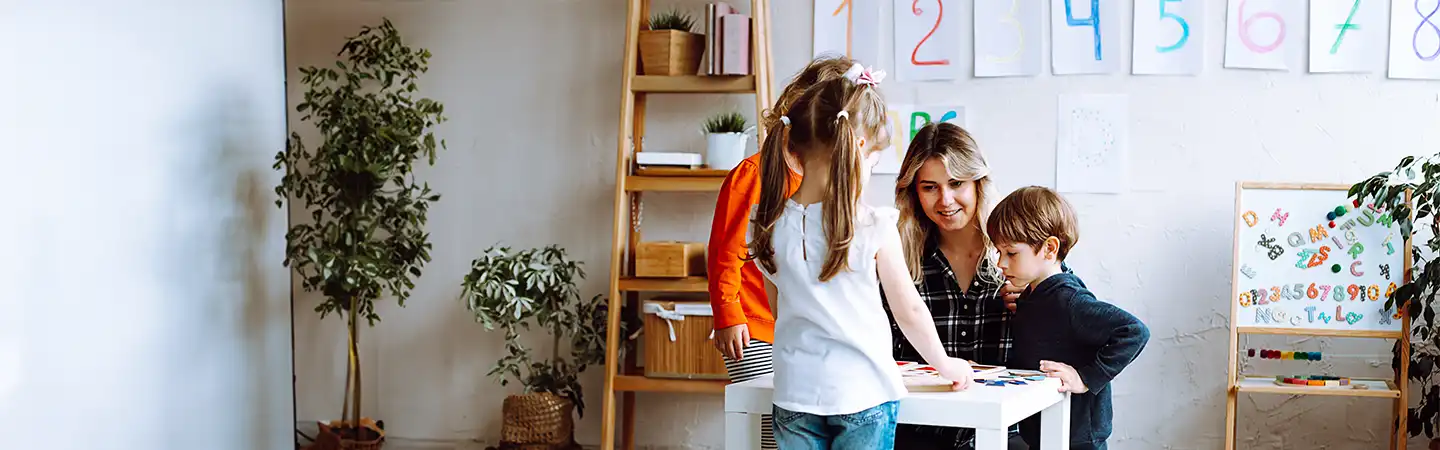 Teacher and three young children gathered around a table in a classroom with educational posters and plants.