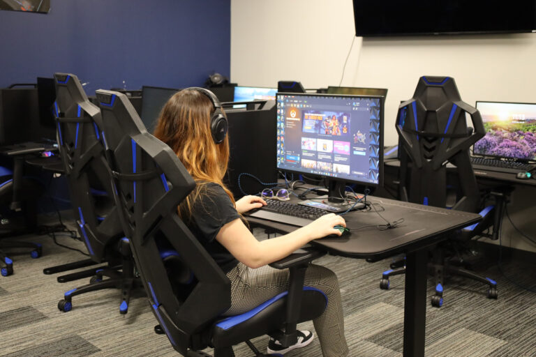 Person wearing headphones using a gaming computer in a room with multiple gaming stations and blue-accented chairs.