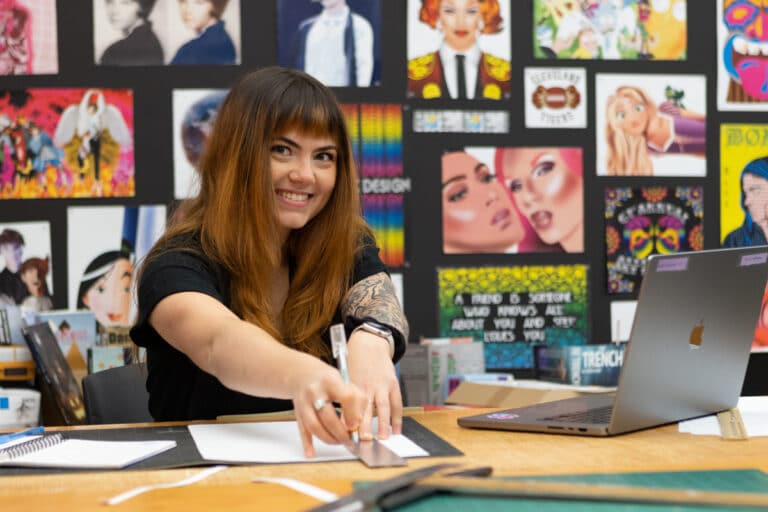 A person smiles while using a ruler at a desk, surrounded by colorful posters and a laptop.