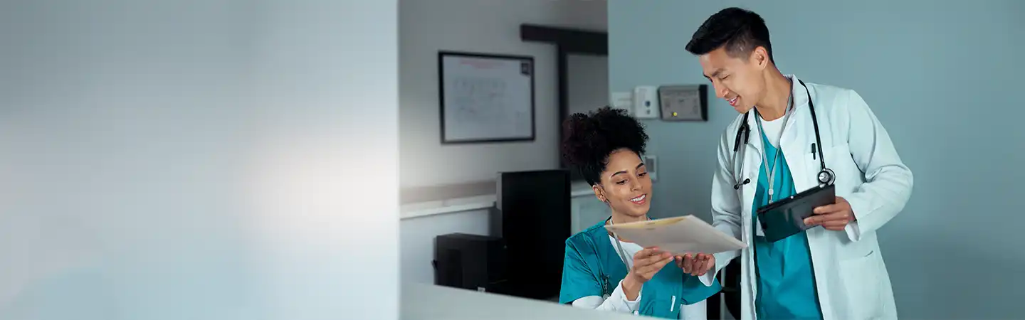 Two healthcare professionals, one seated holding paperwork and the other standing with a tablet, discussing in a medical office.