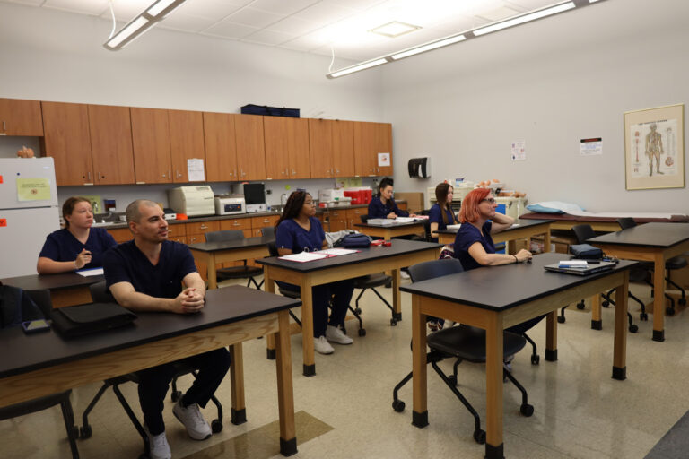Students in navy scrubs seated at tables in a classroom with medical posters and supplies.