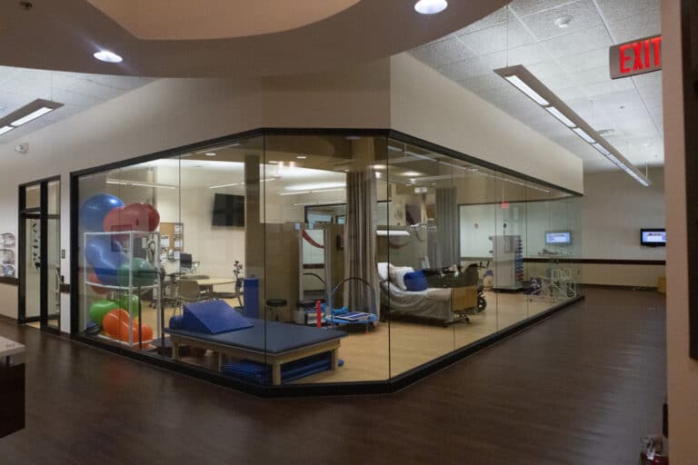 A glass-walled physical therapy room with exercise balls, a treatment bed, and other therapy equipment.