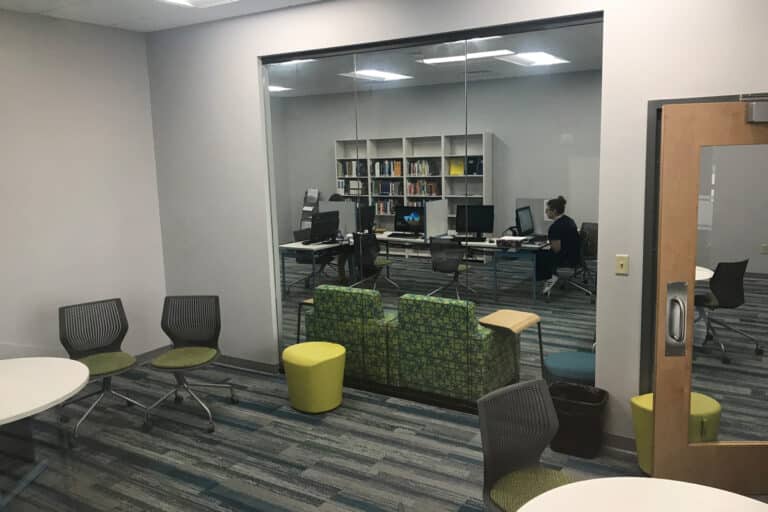 A small office space with glass walls, equipped with desks, computers, and a single person working inside.