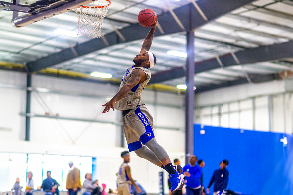 A basketball player in a gym leaps toward the hoop with an outstretched arm, attempting a one-handed slam dunk.