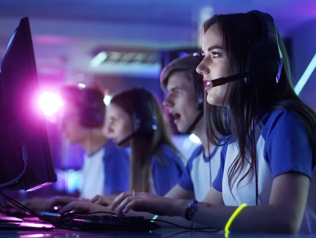 A team of focused young gamers in headsets playing video games on computers in a dimly lit room.